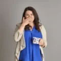 Anna Walton wearing a blue scarf and holding one hand in front of her mouth, the other hand holding a mug