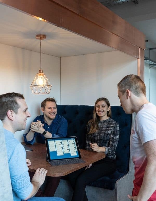 Four colleagues talk at a meeting booth in office, looking at a task board on a laptop