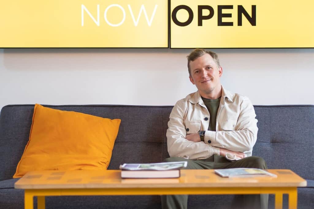 Atomic Smash MD and co-founder Piers Tincknell sitting in front of sign saying "Now Open"