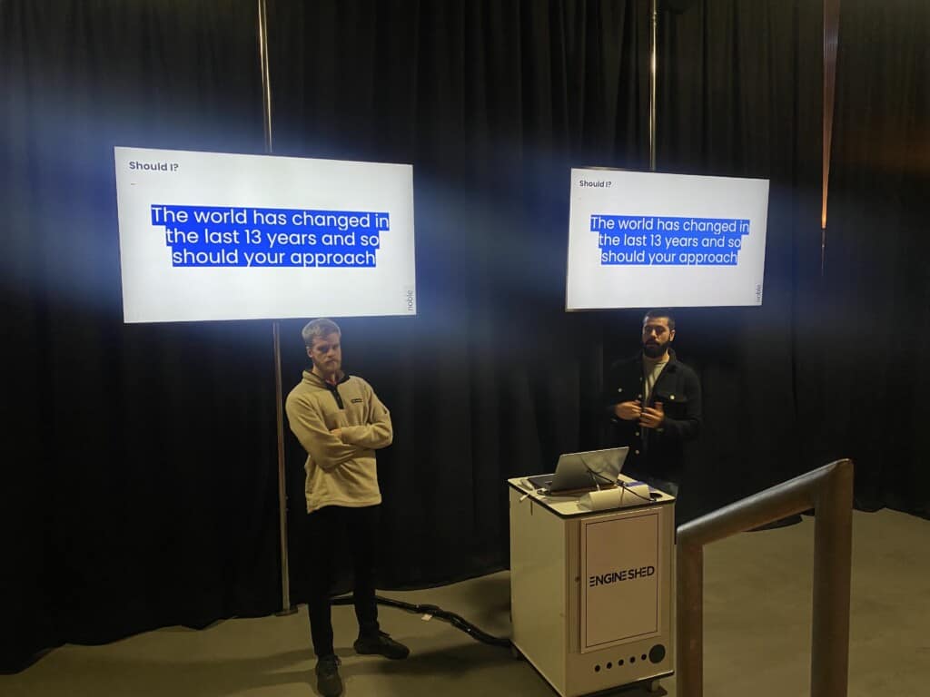 Two men stand in front of screens during presentation at event about how to sell more without costing the earth