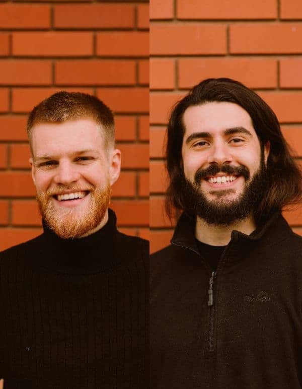 Two men smile wearing black jumpers in front of a brick wall