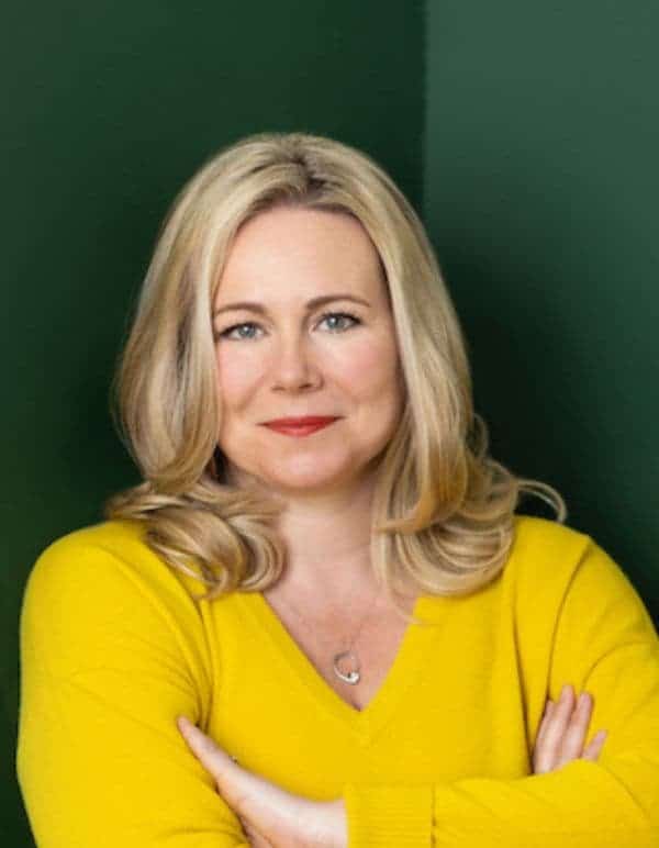 Woman in yellow jumper stands in front of a green wall with her arms crossed