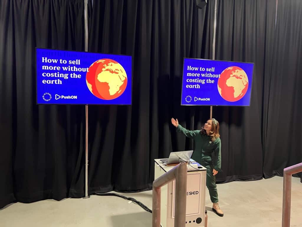 Woman wearing green boilersuit gestures towards screen during presentation about how to sell more without costing the earth
