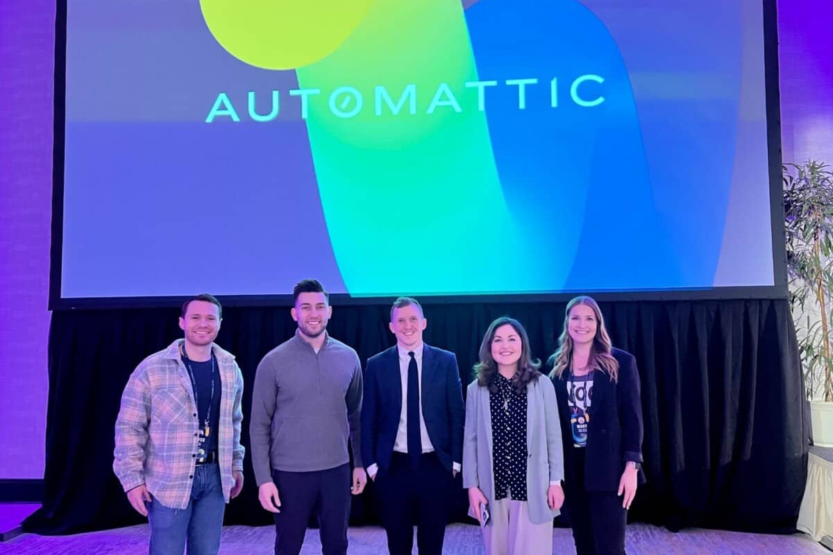 Two team members from Atomic Smash and three team members from WooCommerce stand in front of a screen with the Automattic logo